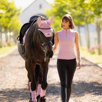 A young woman with a brown bob haircut, wearing a sleeveless pink top and black breeches, walks next to a dark brown horse. The horse wears a pink fly hood, saddle pad, and leg wraps. They are walking down a sunlit gravel path lined with trees, with a building in the background.